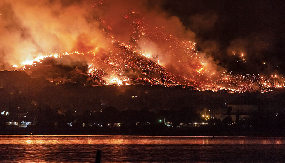 large wildfire on a hillside at night, with flames lighting up the sky and reflections in nearby water