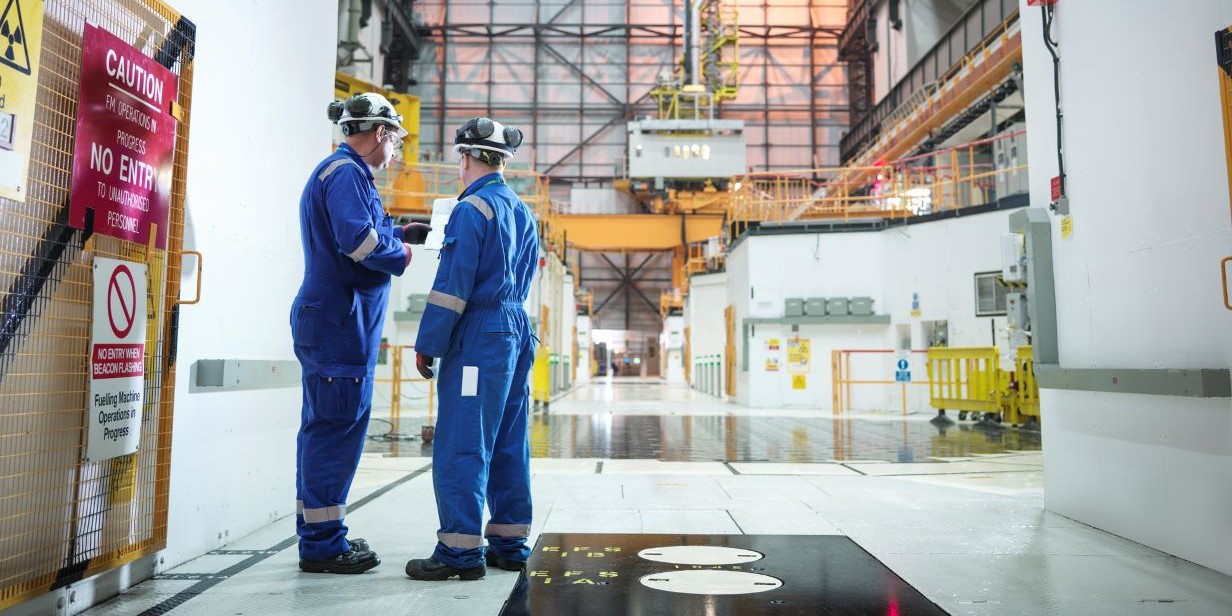 two workers in blue protective suits inside an industrial facility with safety signs and machinery