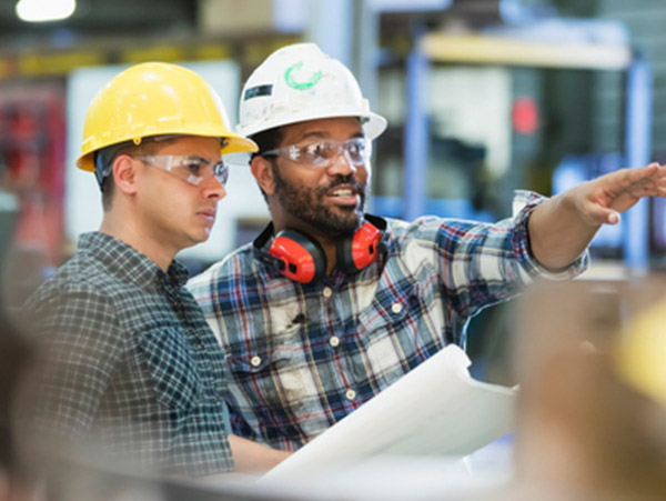 two people in hard hats in an industrial setting, one pointing and one holding a document
